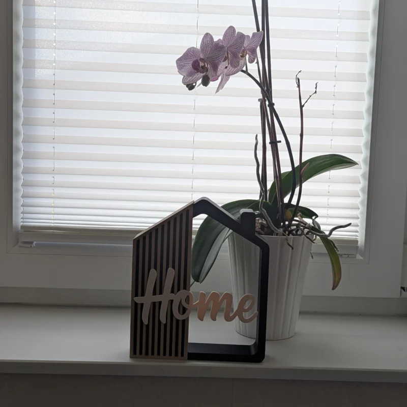 Casita decorativa Home impresa en 3D en negro, arena y crema, en una ventana junto a una orquídea