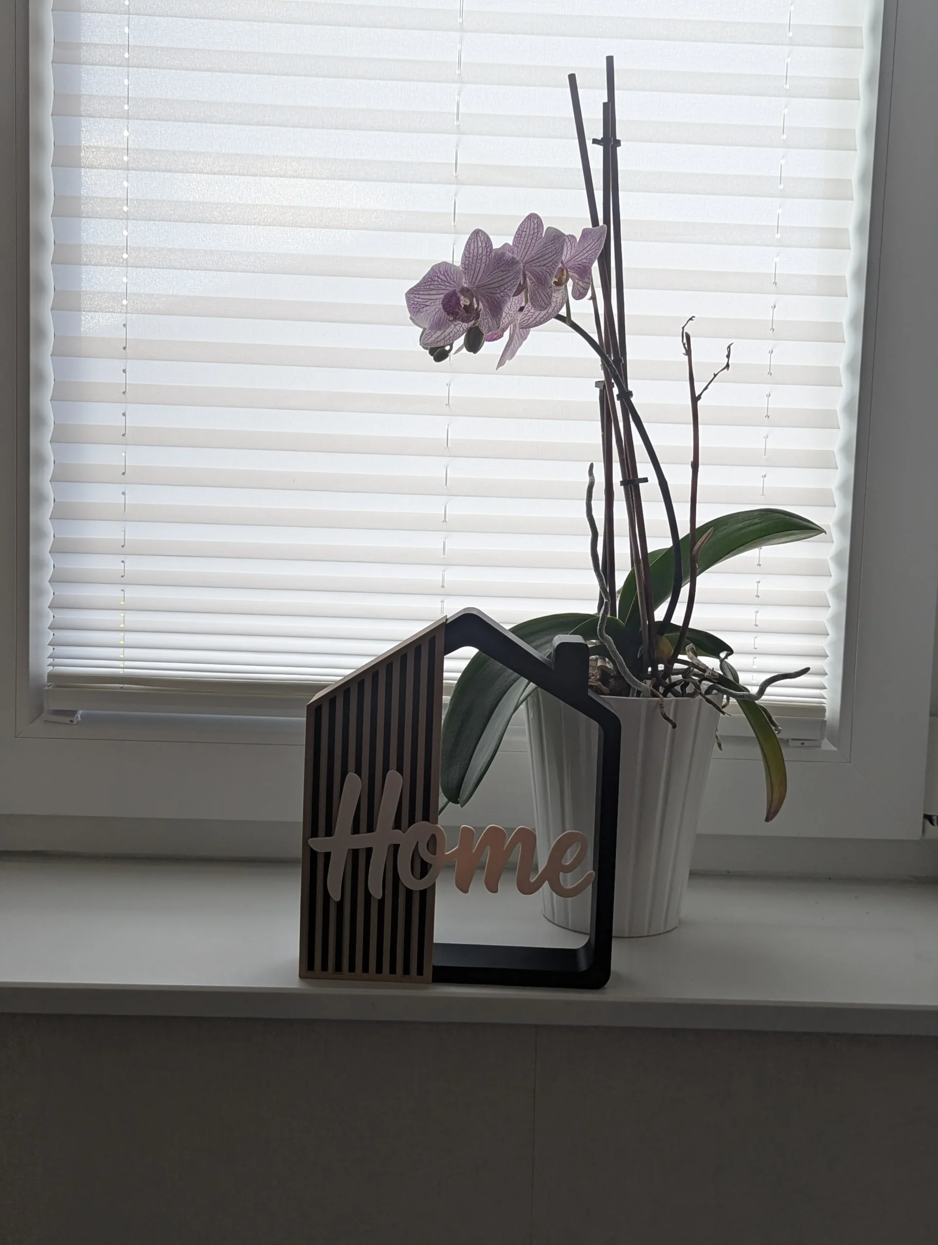 Casita decorativa Home impresa en 3D en negro, arena y crema, en una ventana junto a una orquídea