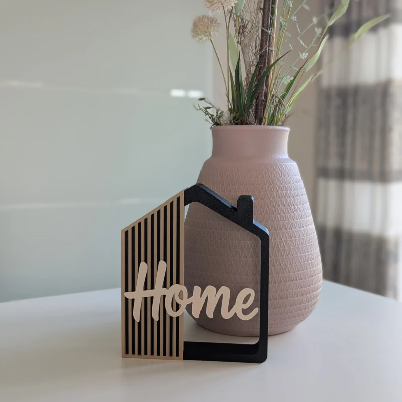 Casita decorativa Home impresa en 3D en negro, arena y crema, junto a un jarrón