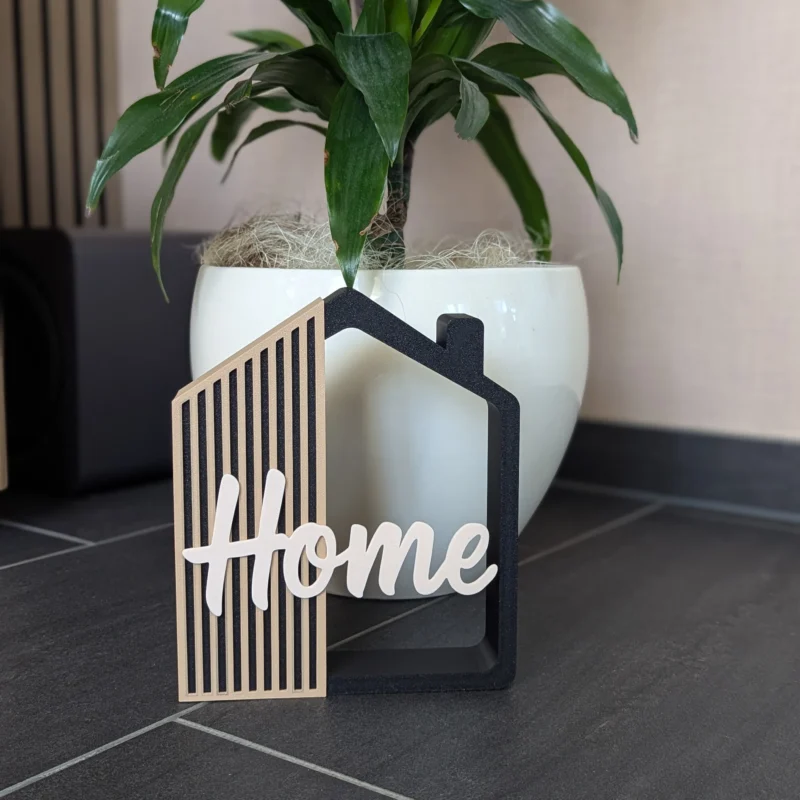 Casita decorativa Home impresa en 3D en negro, arena y crema, junto a una planta