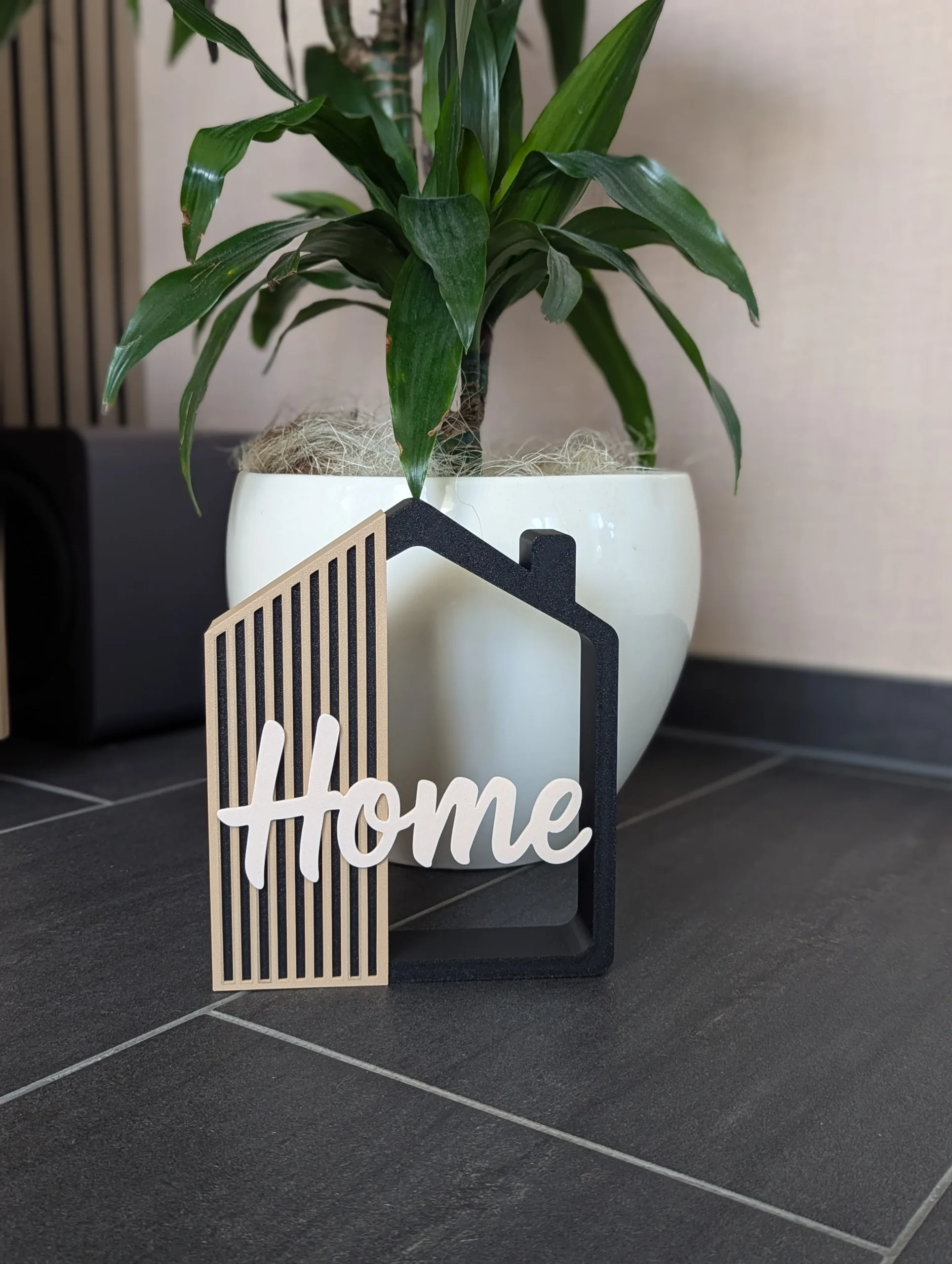 Casita decorativa Home impresa en 3D en negro, arena y crema, junto a una planta