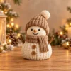 Muñeco de nieve de punto en crema y madera en un ambiente navideño con velas, piñas y luces